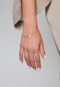 Złota bransoletka o skręconym wzorze, z zawieszką w kształcie serca osadzoną w małych przezroczystych kamieniach. Gładka tekstura, lekka.