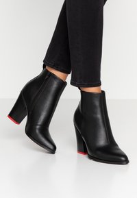 Schwarze Leder-Ankle-Boots mit einer spitzen Zehenpartie und seitlichem Reißverschluss. Sie verfügen über einen klobigen Absatz mit einem roten Akzent an der Sohle. Glatte Textur.