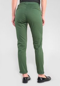 Pantalons verts ajustés avec une texture lisse, présentant des poignets retroussés, deux poches arrière et une taille ajustée. Associés à des chaussures noires.