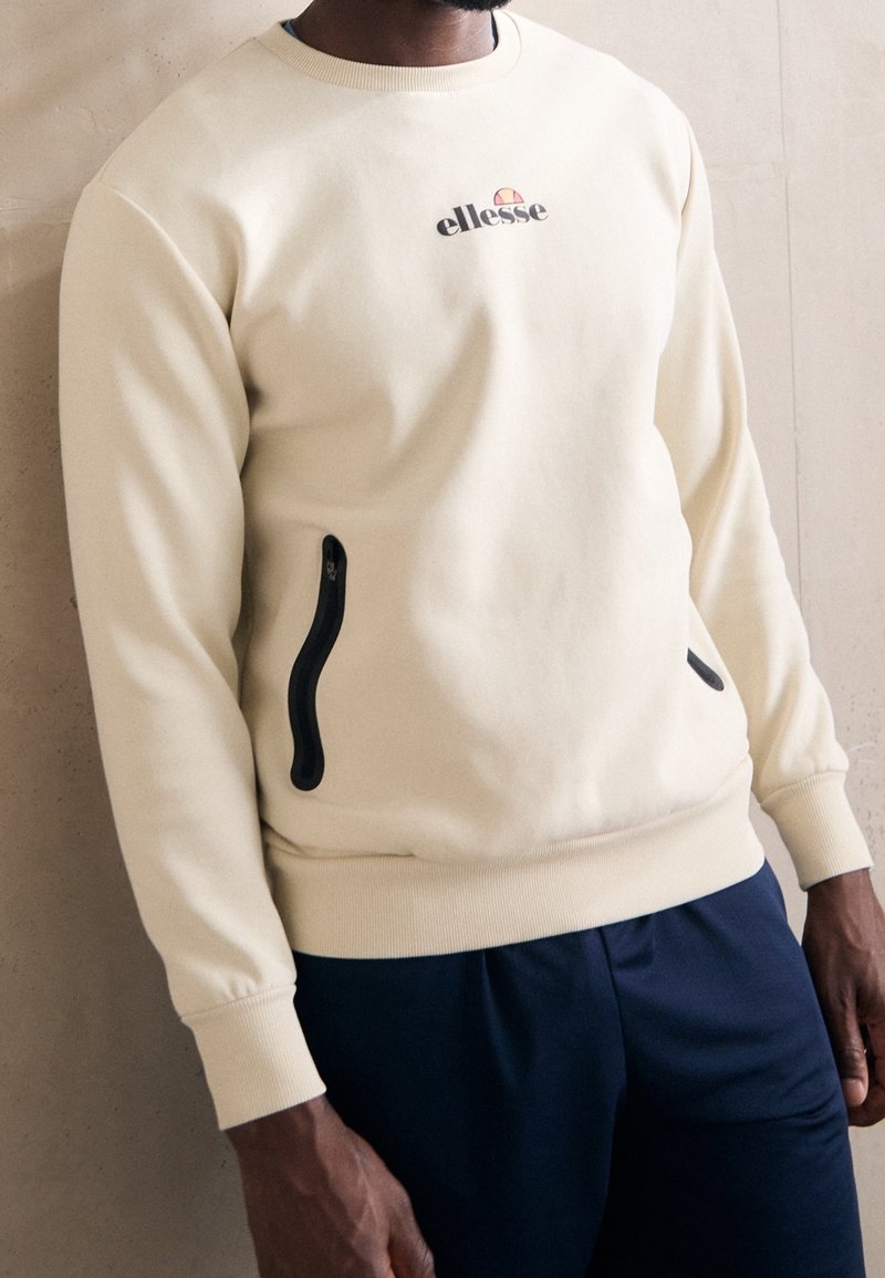 Kremowy sweatshirt z czarnymi kieszeniami na suwak, prążkowanymi mankietami i małym logo na klatce piersiowej. Gładka faktura, casualowy krój.