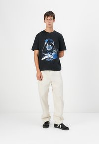 Svart bomull t-shirt med en blå Darth Vader-grafik, ihopparad med löst sittande krämfärgade byxor och svarta sneakers med vita ränder.