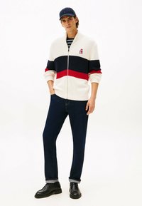 Knitowany sweter z zamkiem w kolorze kremowym, w niebieskie i czerwone paski, zestawiony z ciemnymi dżinsami i czarnymi butami. Posiada logo na klatce piersiowej oraz wysoki kołnierz.