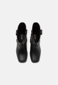 Botines de cuero negro con puntera cuadrada, que cuentan con un detalle de hebilla y una textura suave. Diseño de apertura trasera para un uso fácil.
