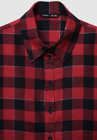 Camicia in flanella a quadri rossi e neri con colletto a punta, bottoni neri e cuciture bianche. Presenta un tessuto texturizzato con un motivo classico.