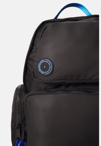 Black nylon backpack with blue accents, featuring a prominent circular patch and a zippered front pocket. Structured design with adjustable straps.