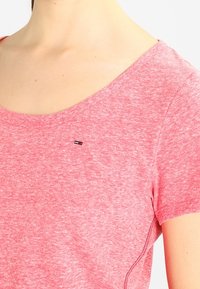 Mujer vistiendo una camiseta corta de manga con un tono rosa jaspeado, un amplio escote redondeado y un pequeño logo rectangular en el pecho.