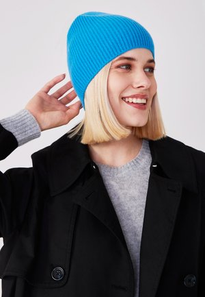 Cappello a maglia a coste blu brillante indossato con un cappotto nero e un maglione grigio. Caratterizzato da una vestibilità aderente e una superficie liscia.