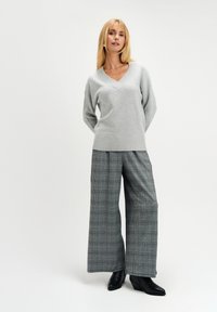 Jasnoszary sweter w serek z prążkowanymi krawędziami, w połączeniu z szerokimi spodniami w czarno-białą kratę. Modelka nosi czarne botki na obcasie.