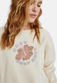 Sudadera de color beige claro con un gran gráfico de hibisco rosa y marrón, junto con el texto "ROXY" y "SURF DAYS" en negrita. Tela suave y texturizada.