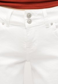 Valge teksapükste lähedalt võetud pilt, millel on topelt hõbedased nupud ja vööaasad, kantud punase soonikkoega pluusiga.