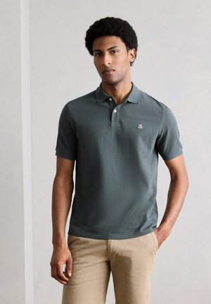 Young man wearing a dark green short-sleeve polo shirt and beige pants, standing against a plain light background with one hand in pocket.