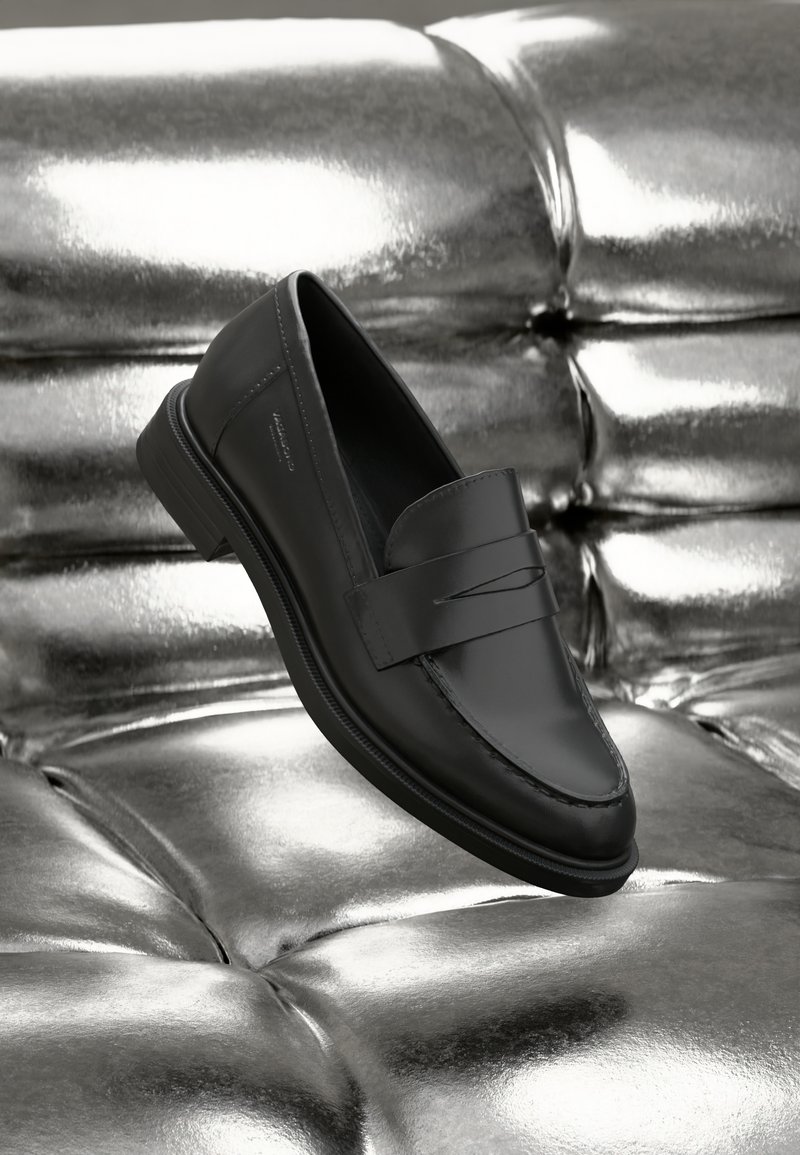 Black leather loafers with a smooth finish, featuring a stitched detail and a raised strip across the front, resting on a metallic surface.