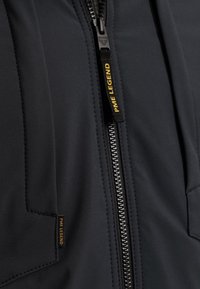 Chaqueta negra con textura suave, que cuenta con una cremallera plateada, bolsillos laterales y una etiqueta negra con "PME LEGEND" en amarillo.