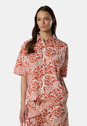 Donna con lunghi capelli scuri che indossa un completo largo abbinato di camicia e pantaloni con motivo floreale arancione e bianco, in piedi con una mano in tasca.