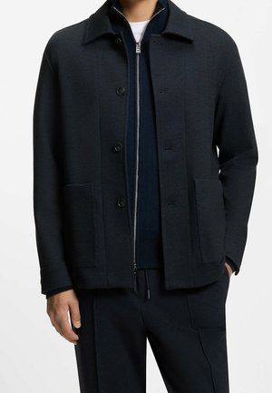 Homme portant une veste noire boutonnée sur un pull bleu marine zippé et un pantalon noir, debout avec une main dans la poche.