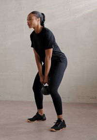 Schwarze Kettlebell, die von einer Person in einem schwarzen Shirt und Leggings gehalten wird, die auf einem hellen Boden vor einer glatten, neutralen Wand steht.