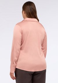 Donna con lunghi capelli castani che indossa una blusa rosa chiaro di seta a maniche lunghe e pantaloni scuri, vista di spalle su uno sfondo neutro.