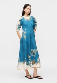 Γυναίκα που φοράει μπλε μίντι φόρεμα με λουλουδάτα σχέδια, φουσκωτά κοντά μανίκια, στέκεται με τα χέρια στις τσέπες και μαύρες σαγιονάρες.