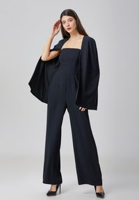 Combinaison noire à jambes larges et corsage sans bretelles, accompagnée d'une cape noire fluide. Le tissu lisse et le design épuré créent une silhouette moderne.