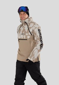 Beige Tarnmuster-Skihjacke mit einer Fronttasche, schwarzen Akzenten und Logodetail am Ärmel; kombiniert mit schwarzen Schneehosen und Handschuhen.