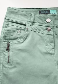 Pantalones de algodón verde claro con un bolsillo con cremallera, acentos de botones de metal y un sutil acabado desgastado alrededor de los bordes del bolsillo.