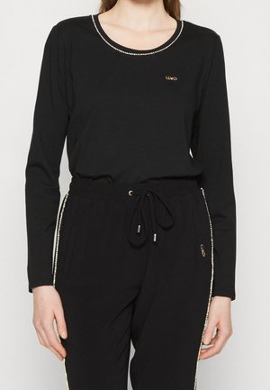 Mujer vistiendo un conjunto negro de top de manga larga y pantalones con adorno de pedrería a lo largo del escote y los lados de los pantalones, manos relajadas a los costados.