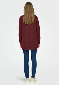 Pull en tricot bordeaux à manches longues, coupe loose et ourlet côtelé, associé à un jean en denim bleu et des baskets beiges.