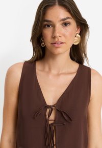 Καφέ αμάνικη μπλούζα με βαθύ V-λαιμόκοψη και δέσιμο μπροστά, φτιαγμένη από ελαφρύ ύφασμα. Φορά μεγάλα χρυσά σκουλαρίκια με σπειροειδές σχέδιο.