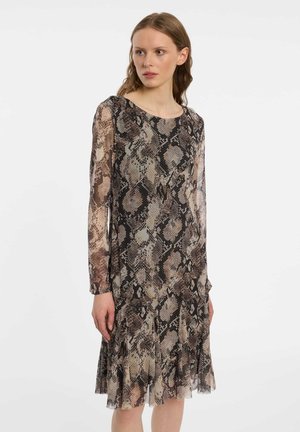 Young woman with light skin and brown hair wears a long-sleeve, knee-length dress with a brown and black snake print pattern.