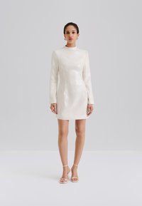 Biele krátke šaty s dlhým rukávom, textúrovaná trblietavá látka, rovný lem a vysoký golier. Doplnkové béžové sandále na remienky.