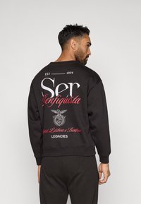 Fekete pulóver, amelynek hátulján nagy fehér és piros felirat található. Dekoratív emblémája egy sas mintát tartalmaz. Puha tapintású, klasszikus fazonú.