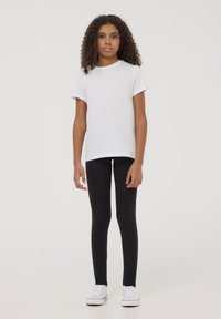 Biała bawełniana koszulka z krótkim rękawem, zestawiona z czarnymi legginsami. Modelka w białych sneakersach, stojąca na jednolitym tle.