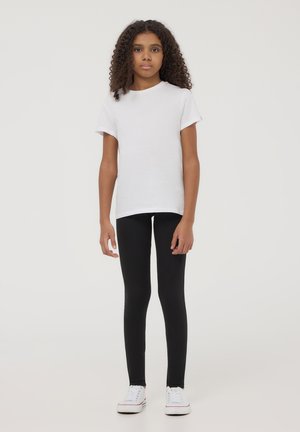 T-shirt bianca in cotone a maniche corte, abbinata a leggings neri. Modello che indossa sneakers bianche, in piedi su uno sfondo neutro.