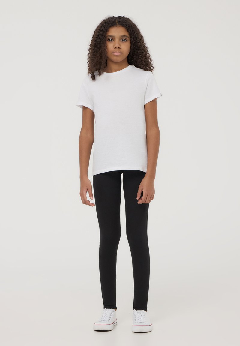 Biała bawełniana koszulka z krótkim rękawem, zestawiona z czarnymi legginsami. Modelka w białych sneakersach, stojąca na jednolitym tle.