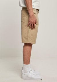 Beige cargo šortky s bočními kapsami, vyrobené z bavlny. Nosit s bílými teniskami a ponožkami, viděno z boku na jednolitém pozadí.