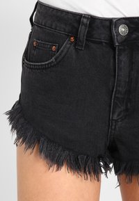 Svarta denimshorts med trasiga sömmar, kopparnitar, fram- och myntfickor, bärs med en vit topp mot en enfärgad bakgrund.