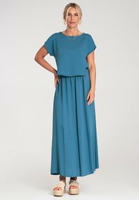 Turkusowa sukienka maxi z luźnym, krótkim rękawem, marszczoną talią i miękką teksturą. Połączona z grubymi, plecionymi sandałami na platformie.