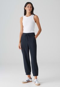 Top sans manches blanc, pantalon de jogging navy ajusté avec cordon de serrage, et baskets blanches avec rayures navy. Tissu lisse et poignets aux chevilles.