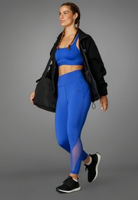 Modrý športový podprsenka a legíny s vysokým pásom a sieťovinovými detailmi, v kombinácii s čiernou bundou na zips a čiernymi teniskami, držiaca čiernu tašku.