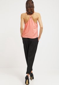 Mujer vestida con un top halter color coral, pantalones negros plisados y tacones altos negros, de pie de espaldas a la cámara sobre un fondo blanco liso.