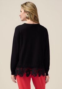 Maglione nero con maniche lunghe, scollatura a coste e orlo in pizzo nero ondulato con motivi floreali. Abbinato a pantaloni rossi.