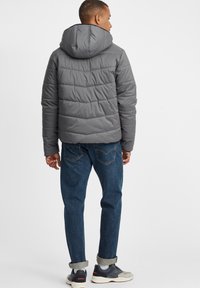 Grå polstret jakke med hette, ribbet krage og horisontal søm. Båret over blå jeans og grå joggesko.