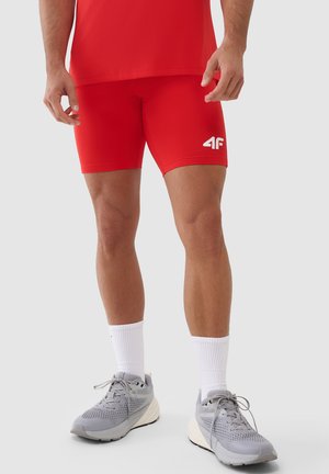 Mężczyzna ubrany w czerwone krótkie spodenki sportowe i pasującą koszulkę, białe skarpetki do kostki oraz szare buty do biegania stoi na jednolitym jasnym tle.