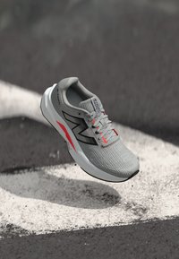 Une chaussure de course gris clair avec un dessus texturé, des accents noirs et néon, un bout arrondi et une semelle rembourrée, apparaissant au-dessus d'une surface peinte.