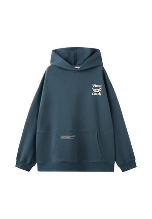 Junger Mann trägt einen dunkelblauen Hoodie mit dem Text "Vinyl Club", beige Shorts und hellbeige Sneakers und steht vor einem schlichten hellen Hintergrund.