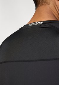 Must sportlik särk, millel on keha järgnev lõige. Omab sujuvat tekstuuri ja peenikesi õmblusi. Selja kaelusel on hõbedane "VENUM" logo.