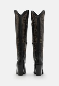 Botas altas de cuero negro con tacón, con punta afilada, que presentan un detallado bordado en tono marrón, un borde ondulado en la parte superior y cremalleras laterales.