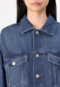 Jeansowa kurtka w jasnoniebieskim odcieniu, z klasycznym kołnierzem, dwiema przednimi kieszeniami z klapkami i srebrnymi zatrzaskami do zapięcia.