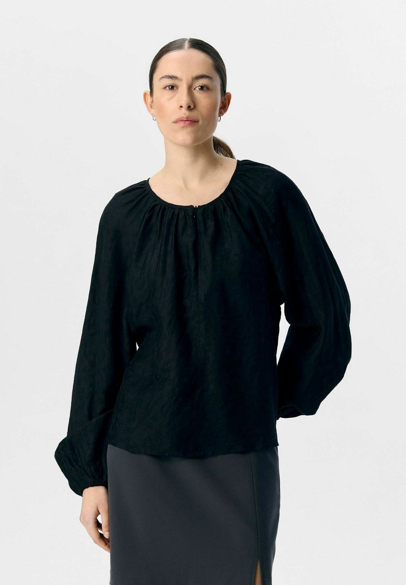 Schwarze Bluse mit langen Ärmeln, gerafftem Rundhalsausschnitt und gerafften Manschetten, aus strukturiertem Stoff. Kombiniert mit einem grauen Rock mit Schlitz.