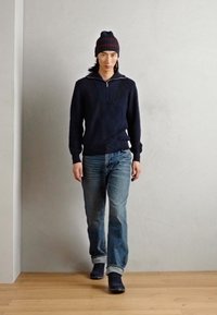 Maglione a maglia navy con zip a un quarto, abbinato a jeans blue chiari e stivali navy. Il modello indossa un berretto in maglia abbinato. Sfondo in legno.